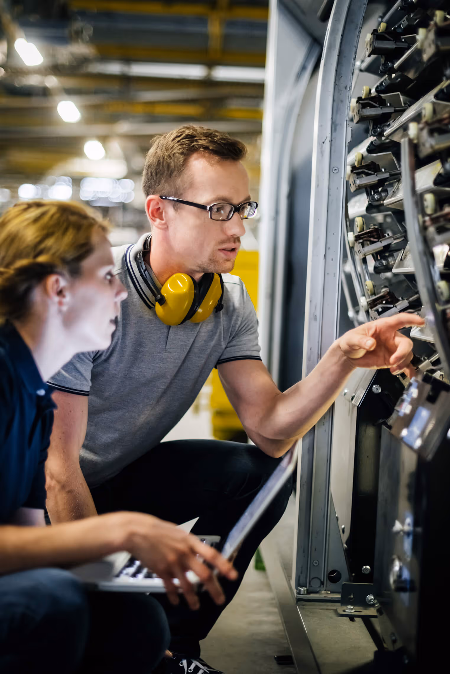 Two factory workers inspecting machinery controls, one pointing and the other holding a laptop.