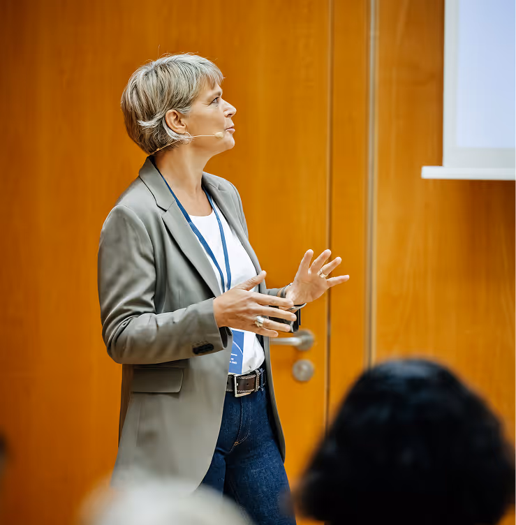 Woman with short hair speaking and gesturing with hands during a presentation in front of a wooden door.