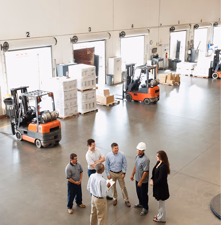 A group of six warehouse workers standing and talking inside a large facility with forklifts and stacked boxes in the background.