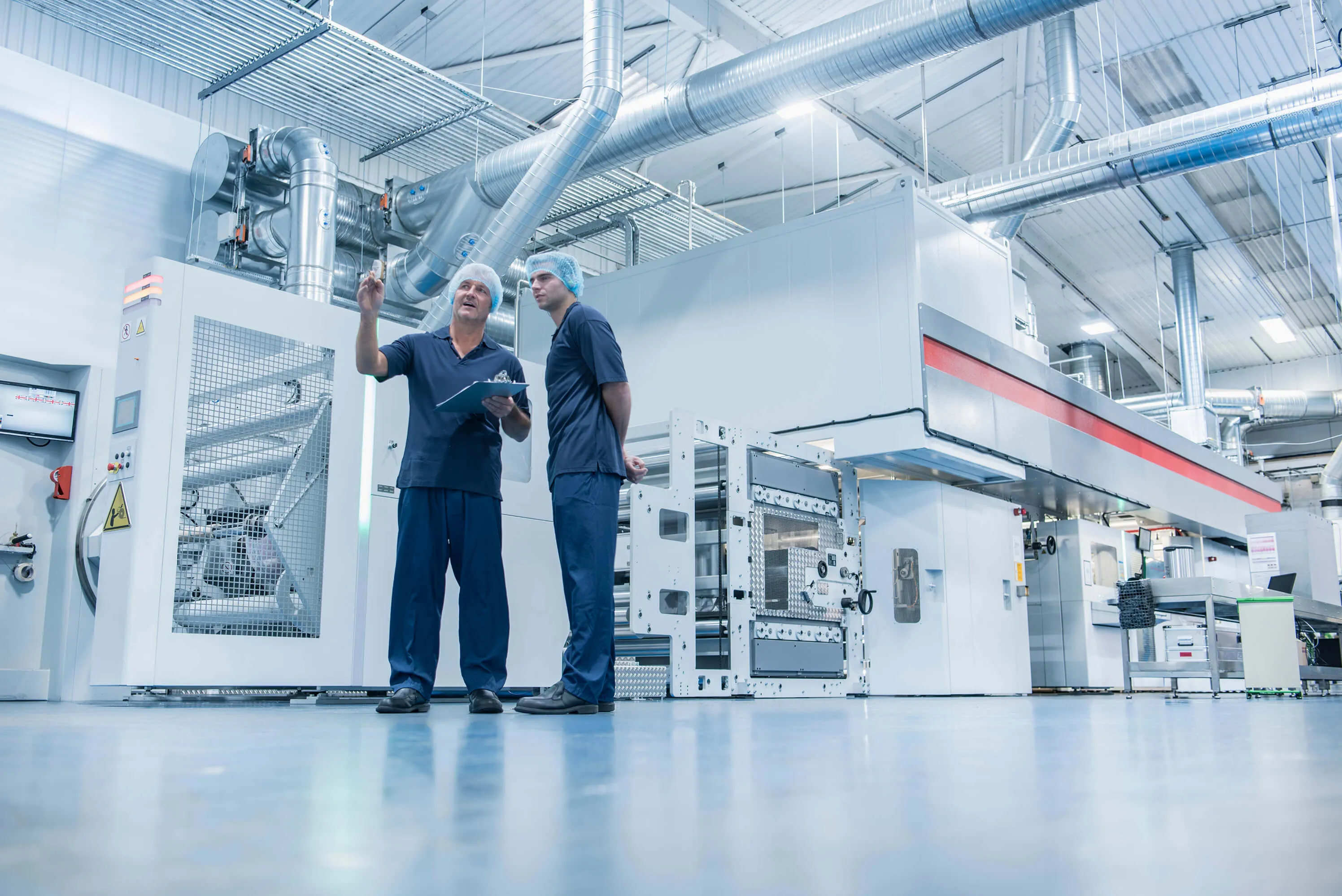 Two factory workers wearing hairnets and navy uniforms discussing near industrial machinery in a clean manufacturing facility.