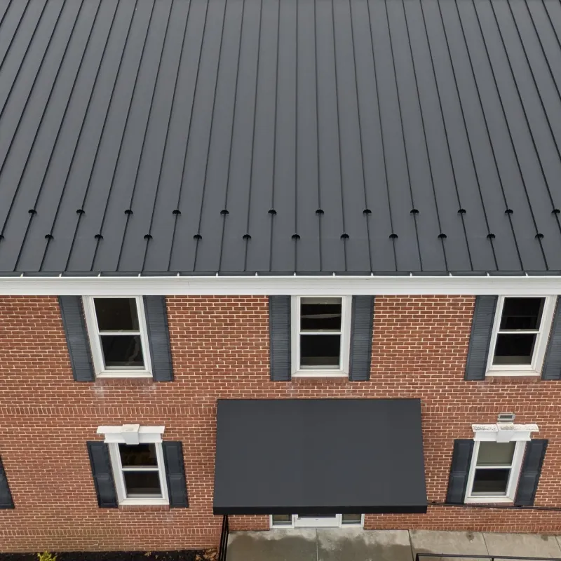Brick building facade with four windows and a gray metal roof featuring vertical ridges and a small black awning above the entrance.