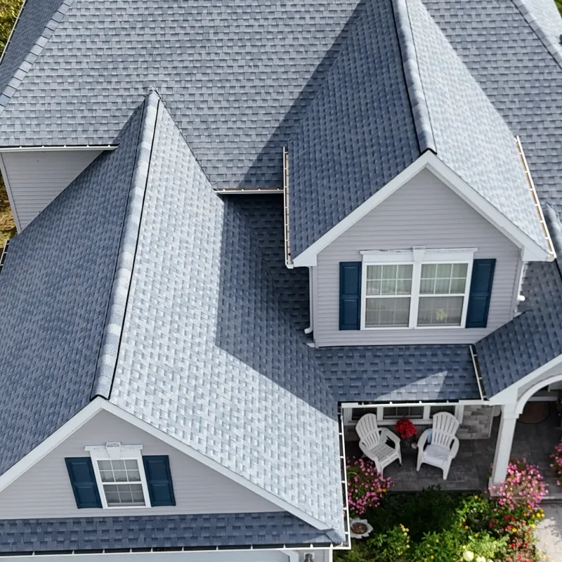 Aerial view of a house with gray asphalt shingle roofing, light gray siding, blue shutters, and a front porch with white chairs and potted flowers.
