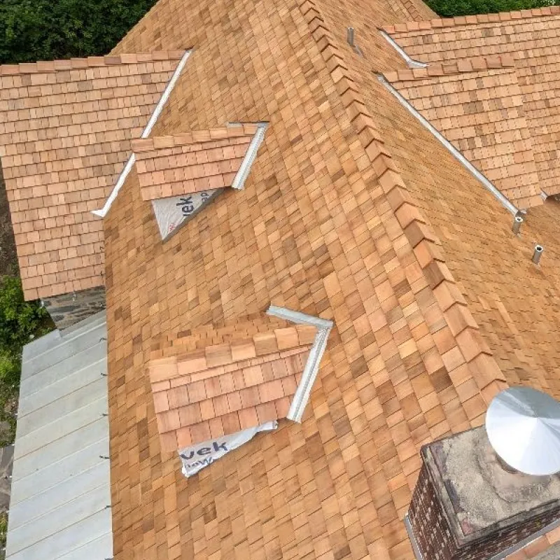 Aerial view of a house roof covered with new cedar wood shingles, featuring two small dormer windows and a brick chimney with a metal cap.