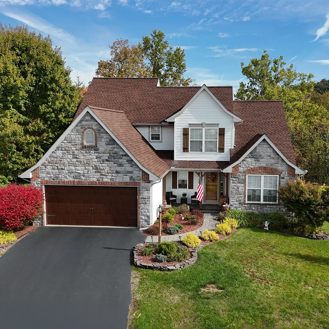 Two-story suburban house with stone and white siding, a brown garage door, a curved walkway, an American flag by the entrance, and well-maintained lawn and shrubs.