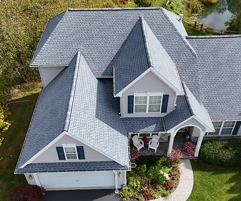 Aerial view of a house with gray asphalt shingle roofing, light gray siding, blue shutters, and a front porch with white chairs and potted flowers.