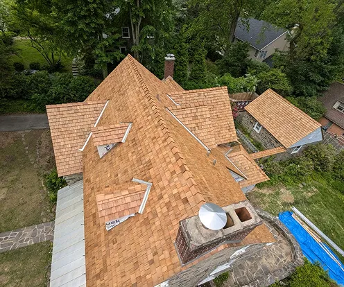 Aerial view of a house roof covered with new cedar wood shingles, featuring two small dormer windows and a brick chimney with a metal cap.