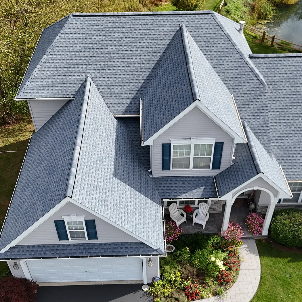 Aerial view of a house with gray asphalt shingle roofing, light gray siding, blue shutters, and a front porch with white chairs and potted flowers.