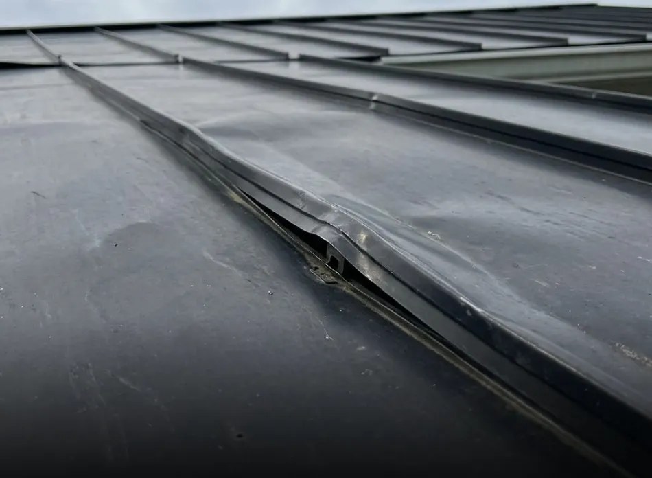 Close-up of a black metal roof panel with a visible bent and damaged seam.