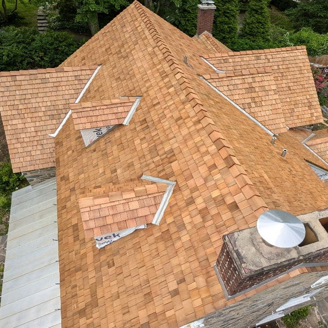 Aerial view of a house roof with newly installed cedar shingles and multiple dormers surrounded by trees.