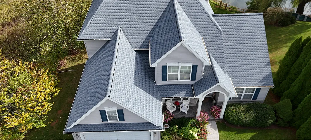 Aerial view of a gray house with blue shutters, a two-chair porch, and surrounding greenery including bushes and trees.