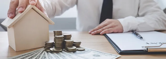 Person in white shirt and black tie holding a wooden house model near stacks of coins and US dollar bills on a desk with a clipboard and pen.