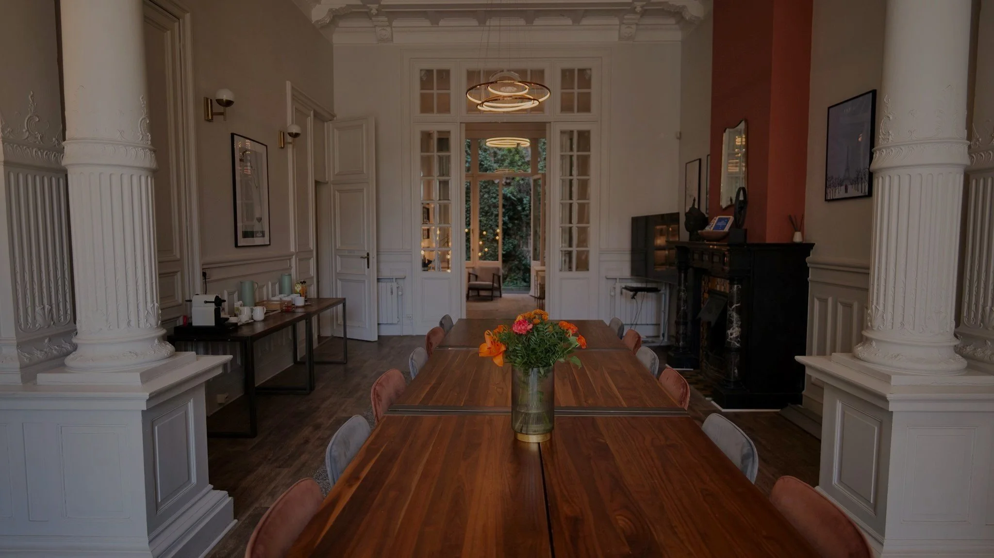 Elegant meeting room with a long wooden table, velvet chairs, flower vase centerpiece, ornate white columns, and glass doors leading to a garden.