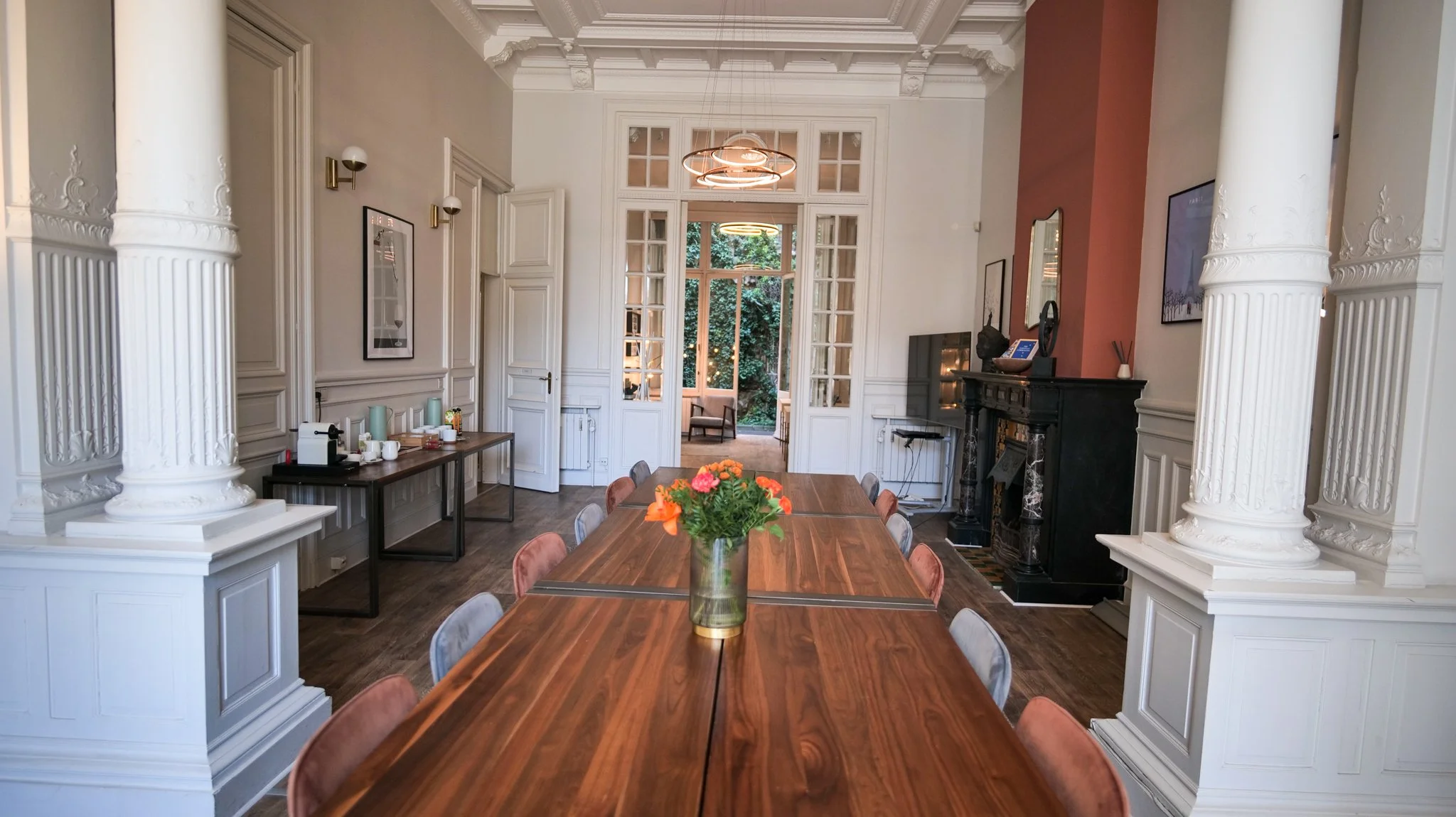 Elegant conference room with a long wooden table surrounded by chairs, a vase with orange flowers, white decorative columns, and a fireplace.