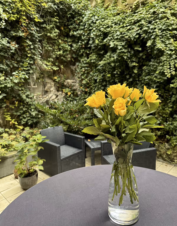 A clear glass vase with a bouquet of yellow roses on a round table in an outdoor garden seating area with black chairs and green ivy-covered walls.