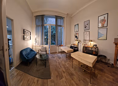 Room with two wooden massage tables, a blue sofa, glass coffee table, and framed anatomical posters on the walls.
