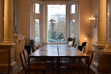 Dining room with a large wooden table surrounded by chairs, featuring a bay window with a view of buildings and trees outside.