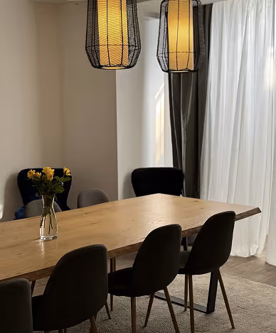 Wooden dining table with six chairs and a glass vase with yellow flowers under two hanging pendant lamps.
