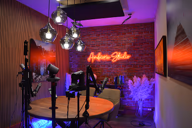 Modern podcast or streaming studio with round wooden table, professional microphones on adjustable arms, hanging industrial lights, neon sign reading 'Ambience Studio', and purple accent lighting.