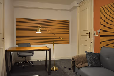 Modern office space with a wooden desk and gray swivel chair, a gold floor lamp, and a gray sofa with pillows and a blanket.