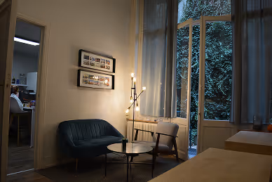 Cozy living room with a dark green sofa, wooden chair, small round coffee table, floor lamp, and large window with sheer curtains.