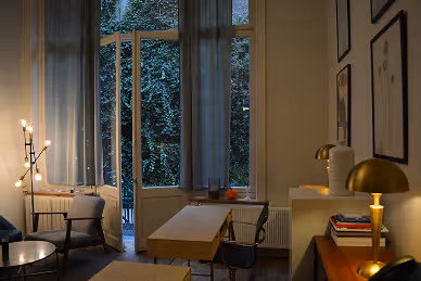 Cozy living room with warm lighting, a coffee table, chairs, books, and an open window with curtains revealing greenery outside.