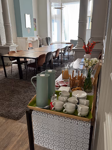 A coffee and tea station with cups, thermoses, juice, and flowers set on a tray in a bright meeting room with tables and chairs.