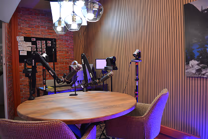 Podcast recording studio with round wooden table, microphones, cameras, and cushioned chairs under hanging lights.