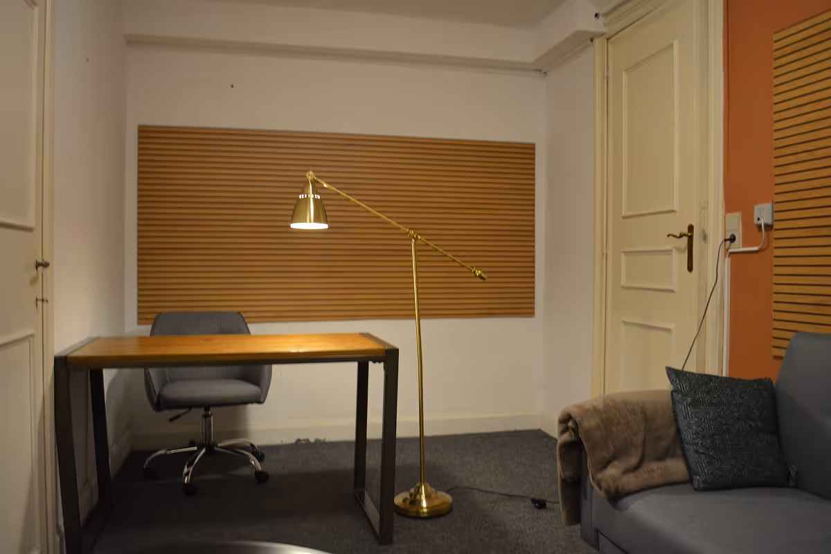 Minimalist office with a wooden desk, gray swivel chair, gold floor lamp, and gray sofa with a pillow and blanket.