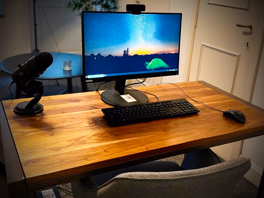 Workspace with a desktop monitor showing a night sky with stars and a green tent, a keyboard, mouse, and microphone on a wooden desk.