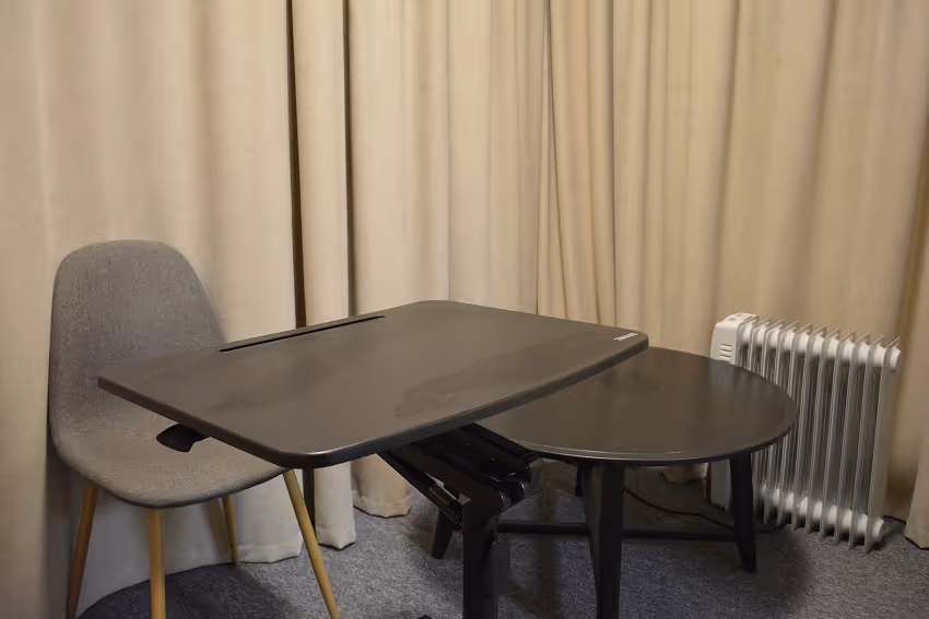 Gray chair with wooden legs next to a small extendable black table and a black round table in a room with beige curtains and a white electric radiator.