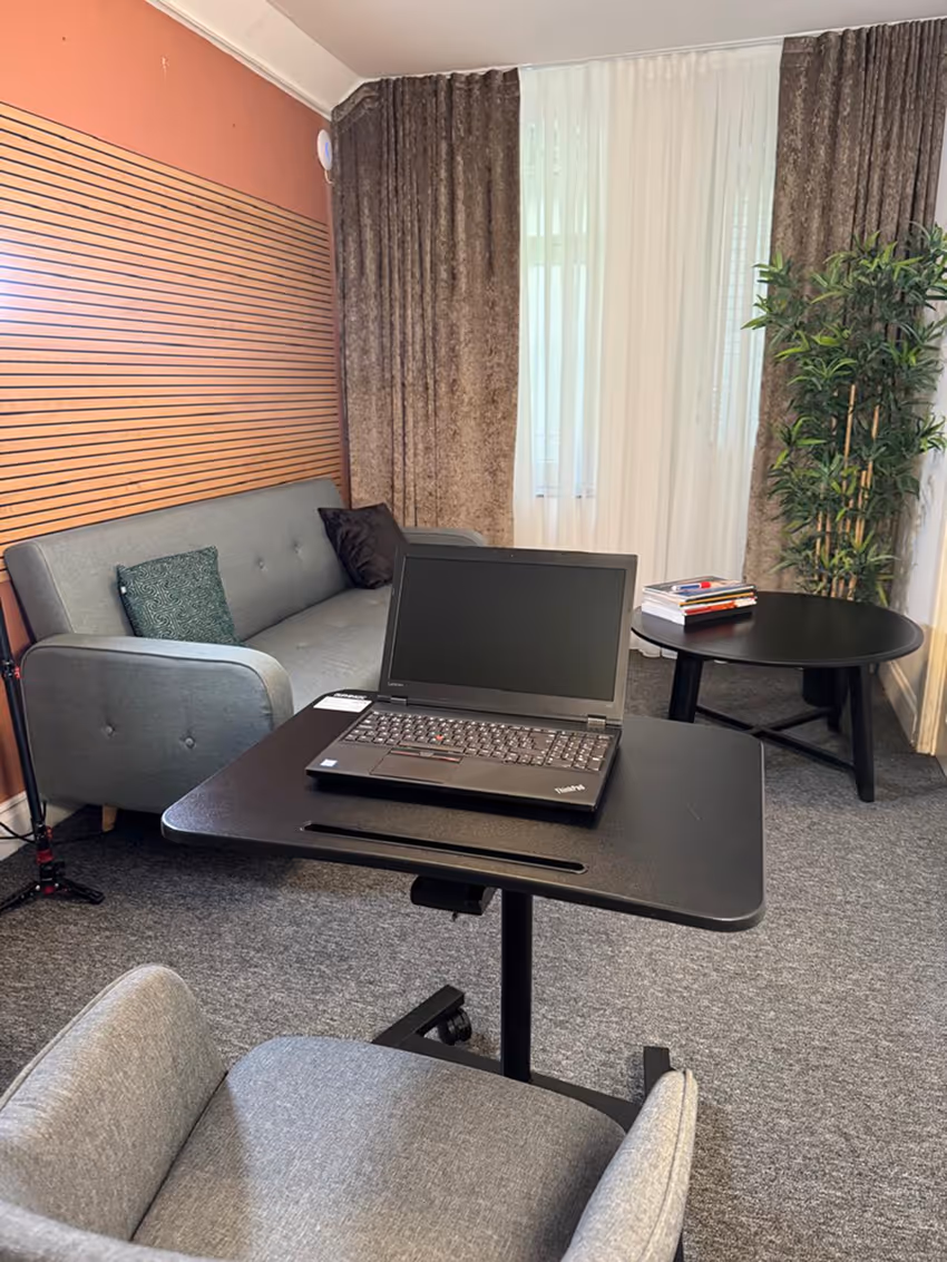 Office space with a black adjustable laptop table holding a closed laptop, a gray upholstered chair in front, a gray sofa with pillows, a round black coffee table with books, and a tall plant near the window with brown curtains.