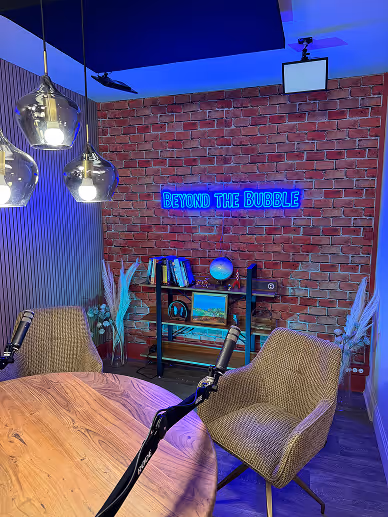 Podcast or interview setup with two wicker chairs, microphones, a wooden table, and a blue neon sign reading 'BEYOND THE BUBBLE' on a brick wall.