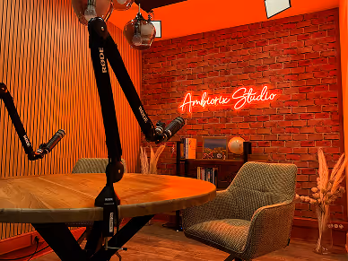 Podcast studio with microphone arms, a round wooden table, an armchair, and a red neon sign reading 'Ambiance Studio' on a brick wall.