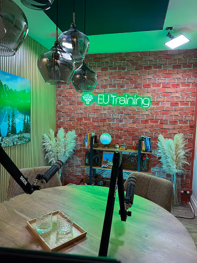 Modern recording studio with round wooden table, microphones, pendant lights, brick wall with EU Training neon sign, and decorative plants.