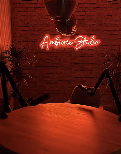 Podcast studio with a round wooden table, two microphones, chairs, and a red neon sign reading 'Ambiorix Studio' on a brick wall.