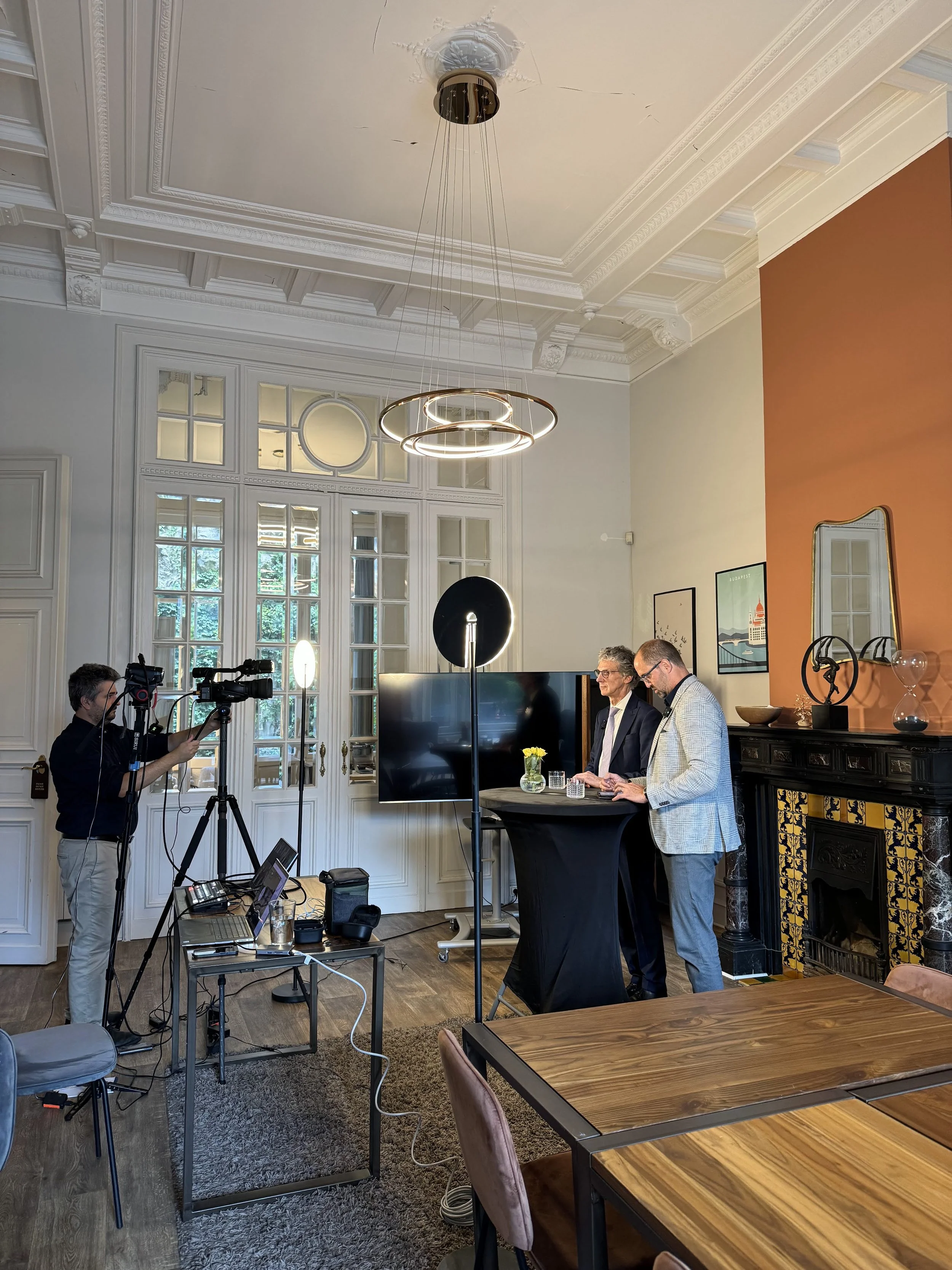 Two men standing behind a round table giving a talk or interview with a cameraman filming them in a modern room with white doors and a decorative fireplace.