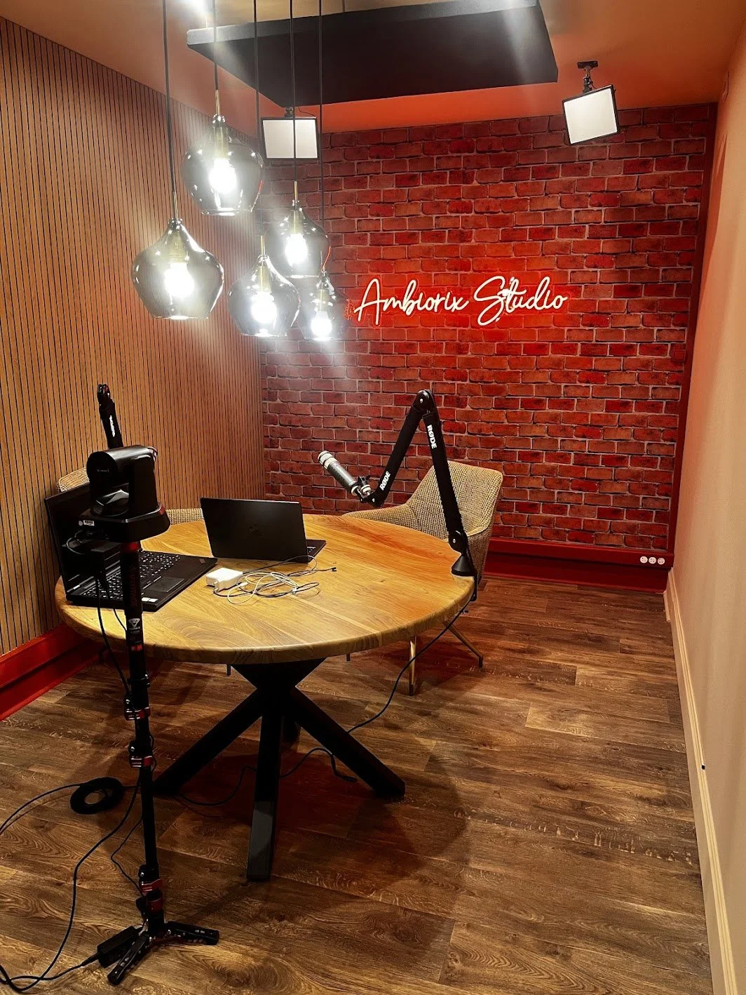 Podcast studio with a round wooden table, two laptops, a microphone on a boom arm, modern hanging lights, and a neon sign reading Ambiorix Studio on a brick wall.