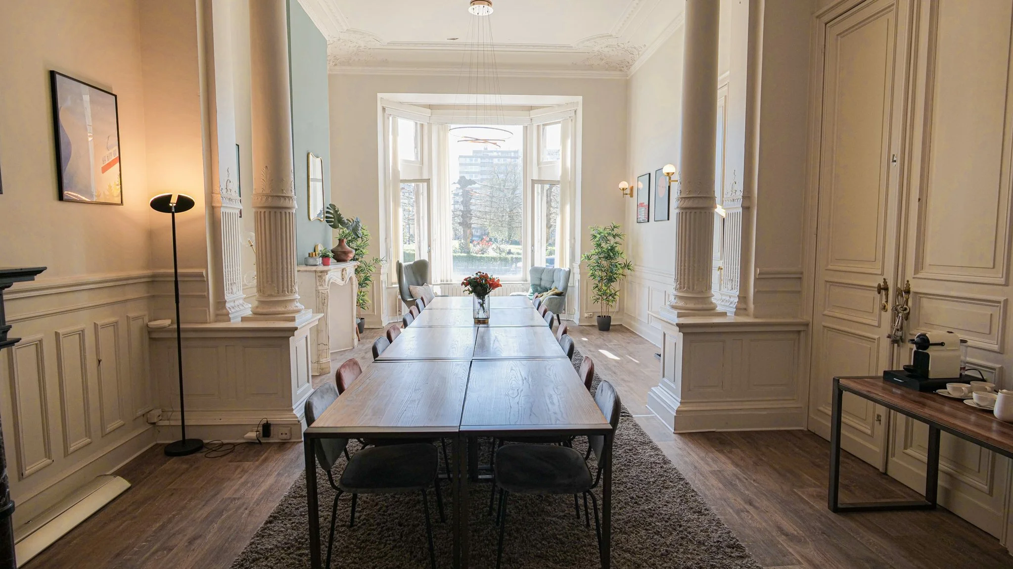 Bright, elegant conference room with a long wooden table, surrounded by chairs, large windows, and decorative columns.