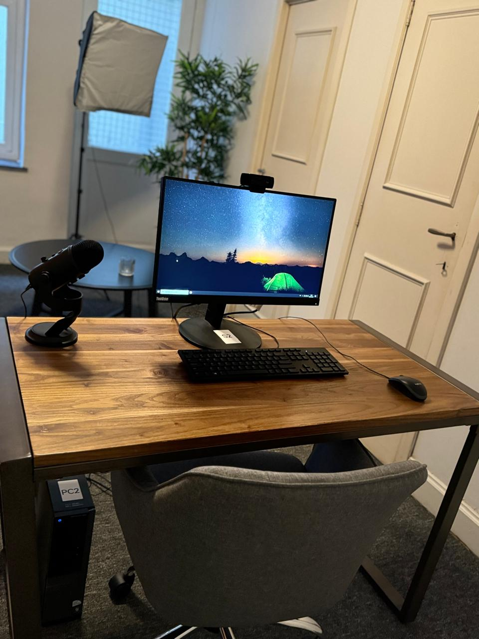 Wooden desk with a computer monitor displaying a starry night sky and tent, black keyboard, mouse, and a microphone on a stand in an office setting.