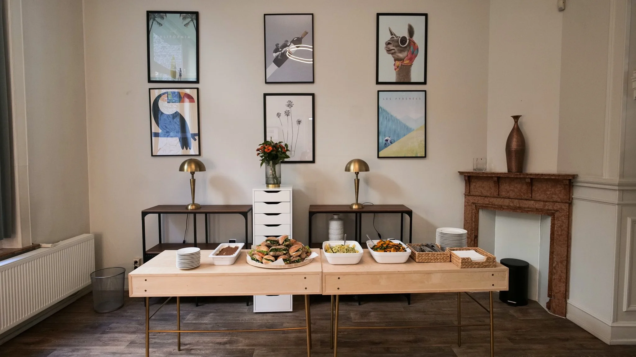 Buffet table with plates, sandwiches, and dishes set in a room with framed art on the wall, two brass lamps, and a decorative fireplace.