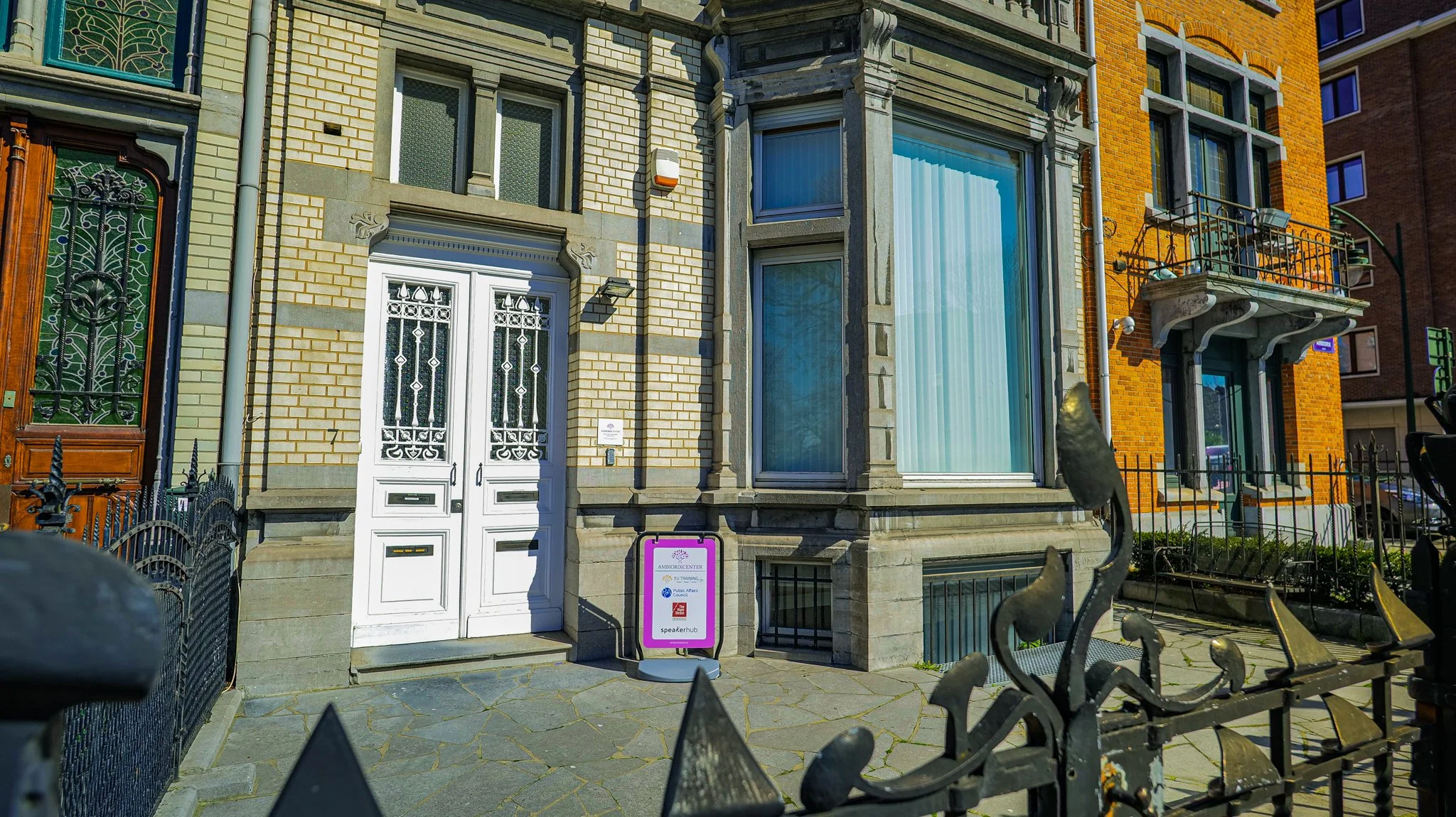 Building entrance with white double doors and a signboard reading Ambassador Center alongside a black wrought iron fence.