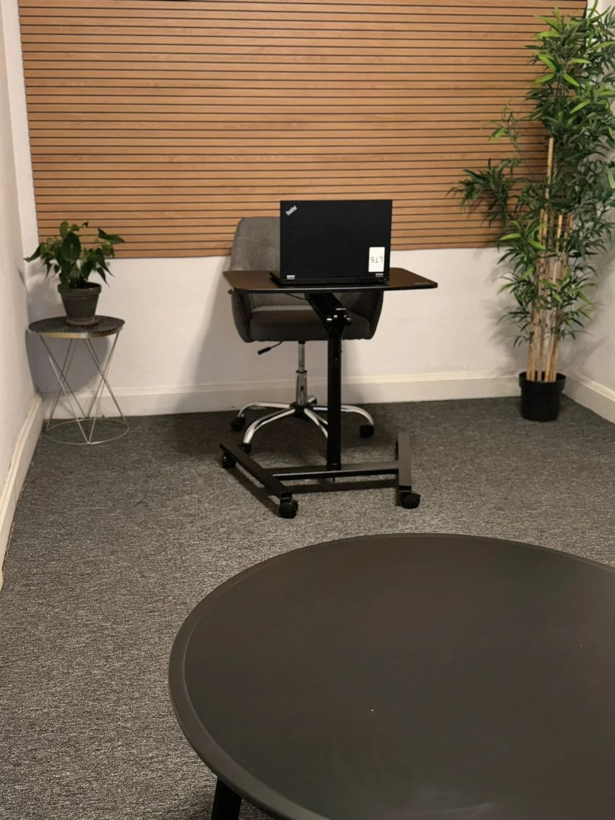 Modern office corner with a laptop on a movable desk, an office chair, potted plants, and a round black table in the foreground.