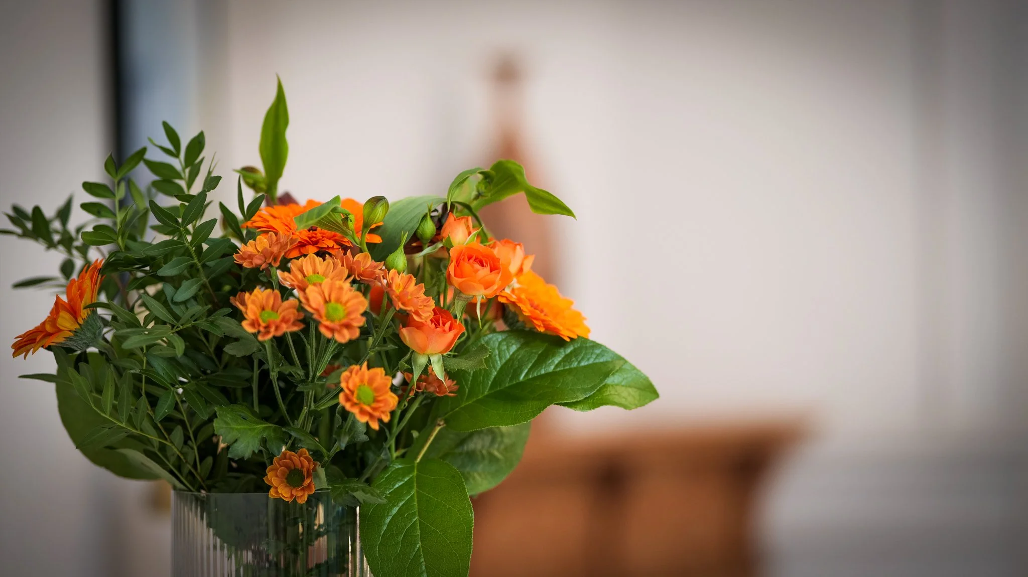 Bouquet of orange flowers and green leaves in a glass vase with a blurred background.
