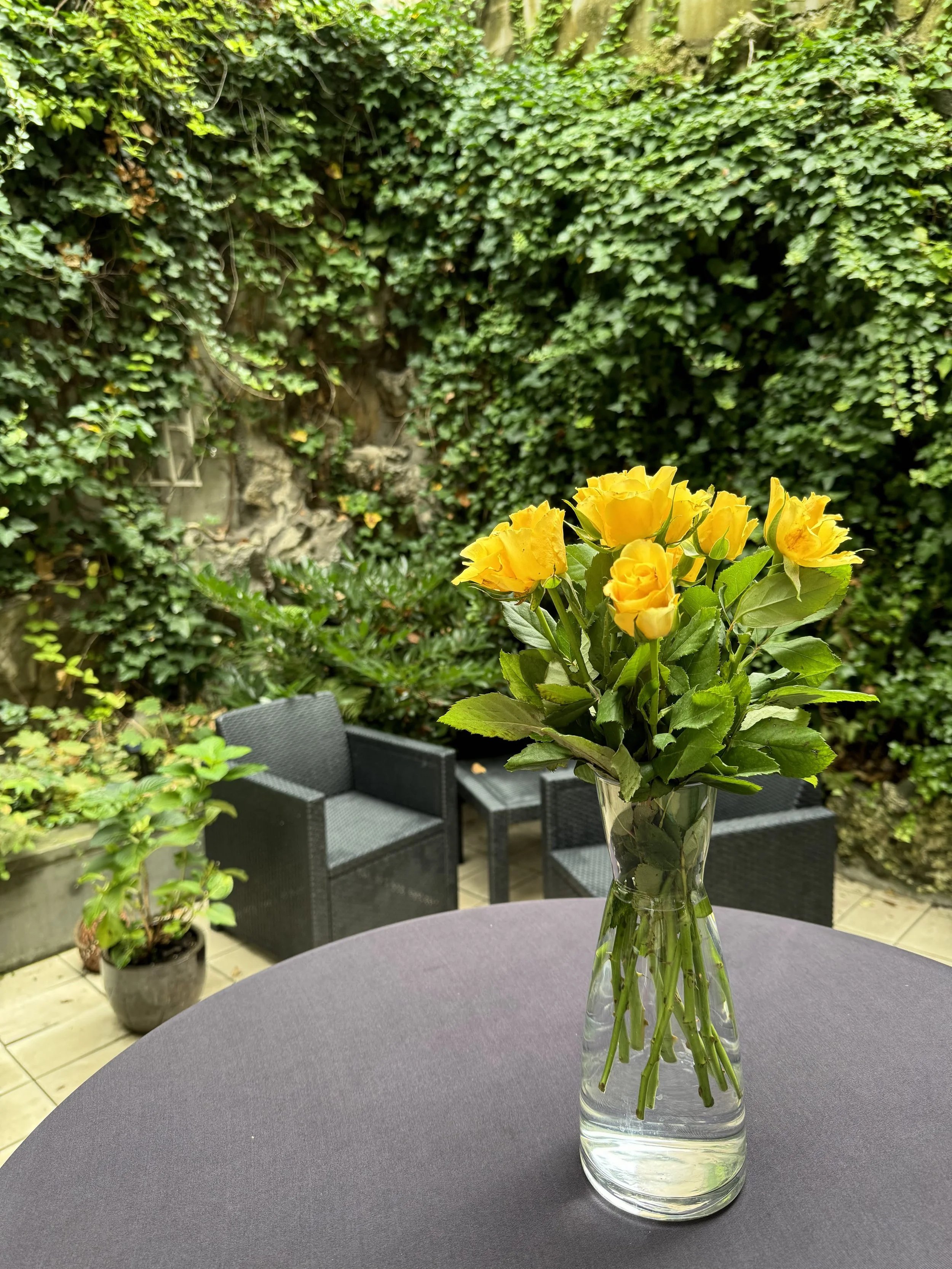 A clear glass vase with a bouquet of yellow roses on a round table in an outdoor garden seating area with black chairs and green ivy-covered walls.