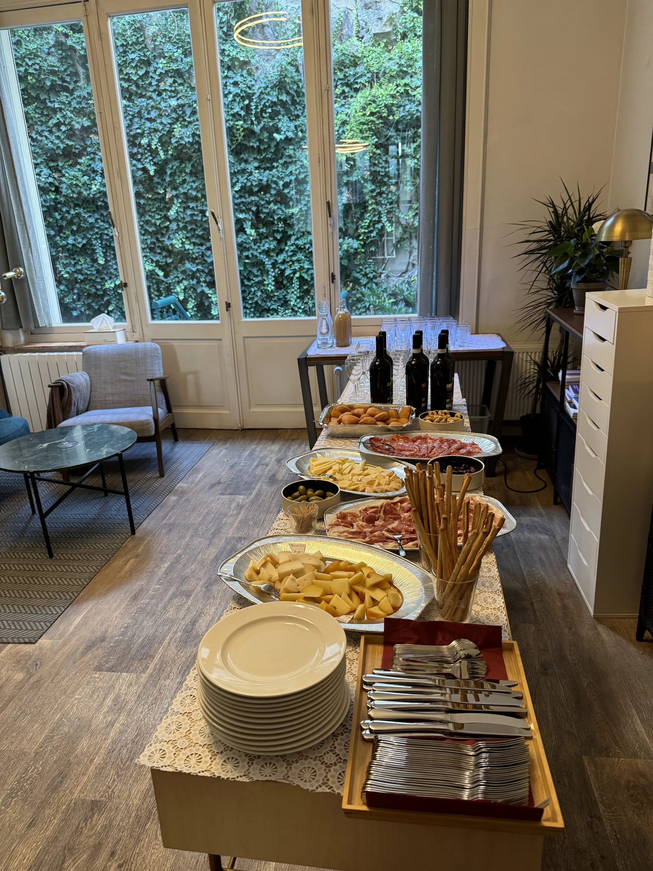 Indoor buffet table with plates, cutlery, breadsticks, assorted cheeses, sliced meats, olives, drinks, and glasses in a room with large windows and green ivy outside.