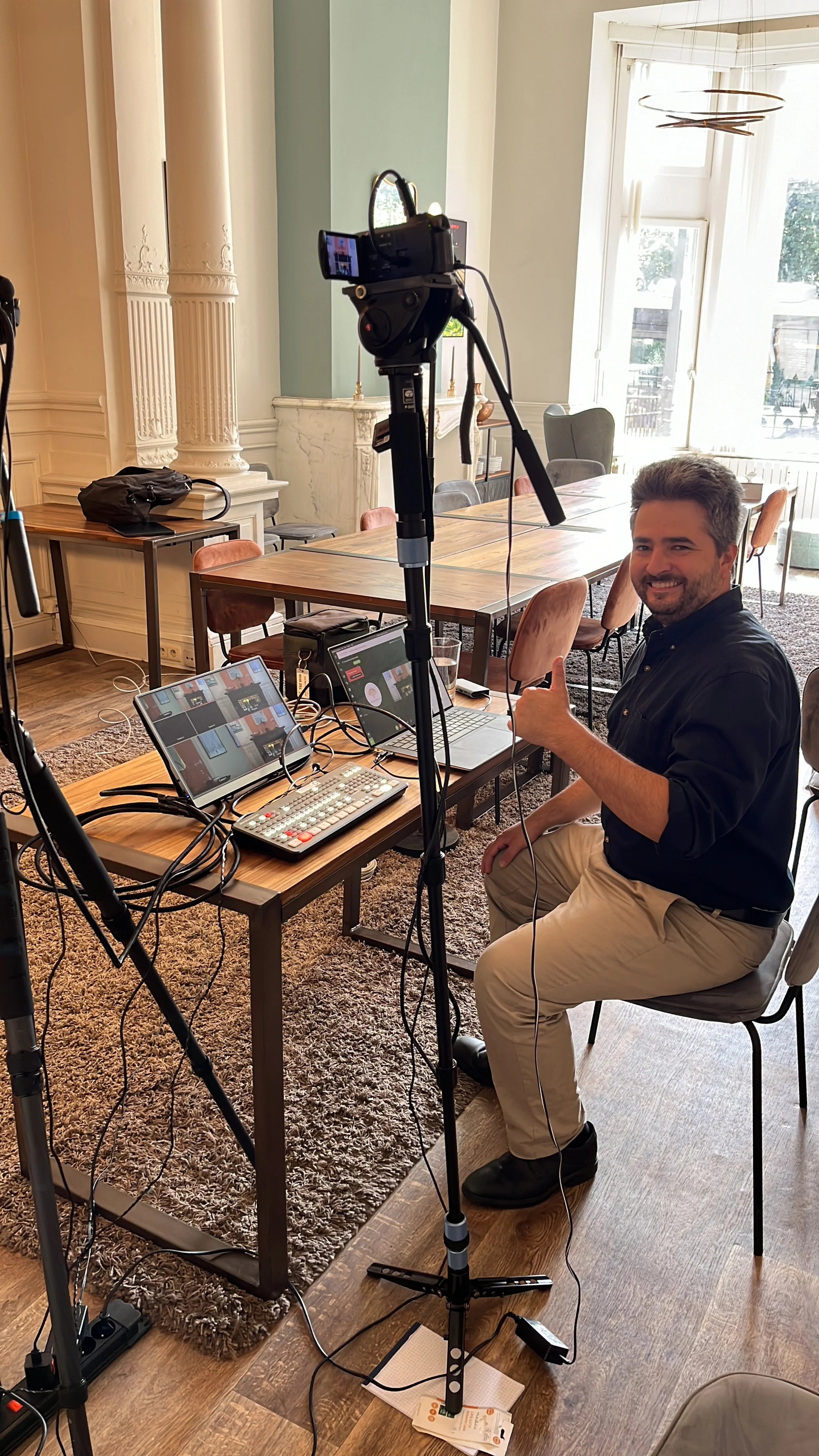 Man sitting on a chair at a wooden table with video production equipment, giving a thumbs-up in a bright room with tall windows and ornate columns.