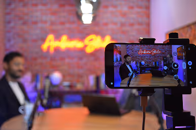 Smartphone on a tripod recording a man speaking in front of a neon sign that reads 'Andrew Solis' against a brick wall.