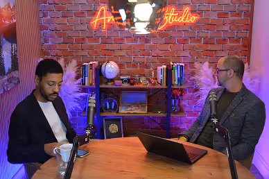 Two men in a podcast studio with microphones, bookshelves, and neon sign reading 'Art Studio' on a brick wall.