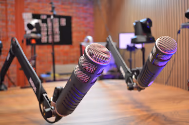 Two professional studio microphones on adjustable arms in a recording or podcast setup with a blurred background of equipment and wooden wall.