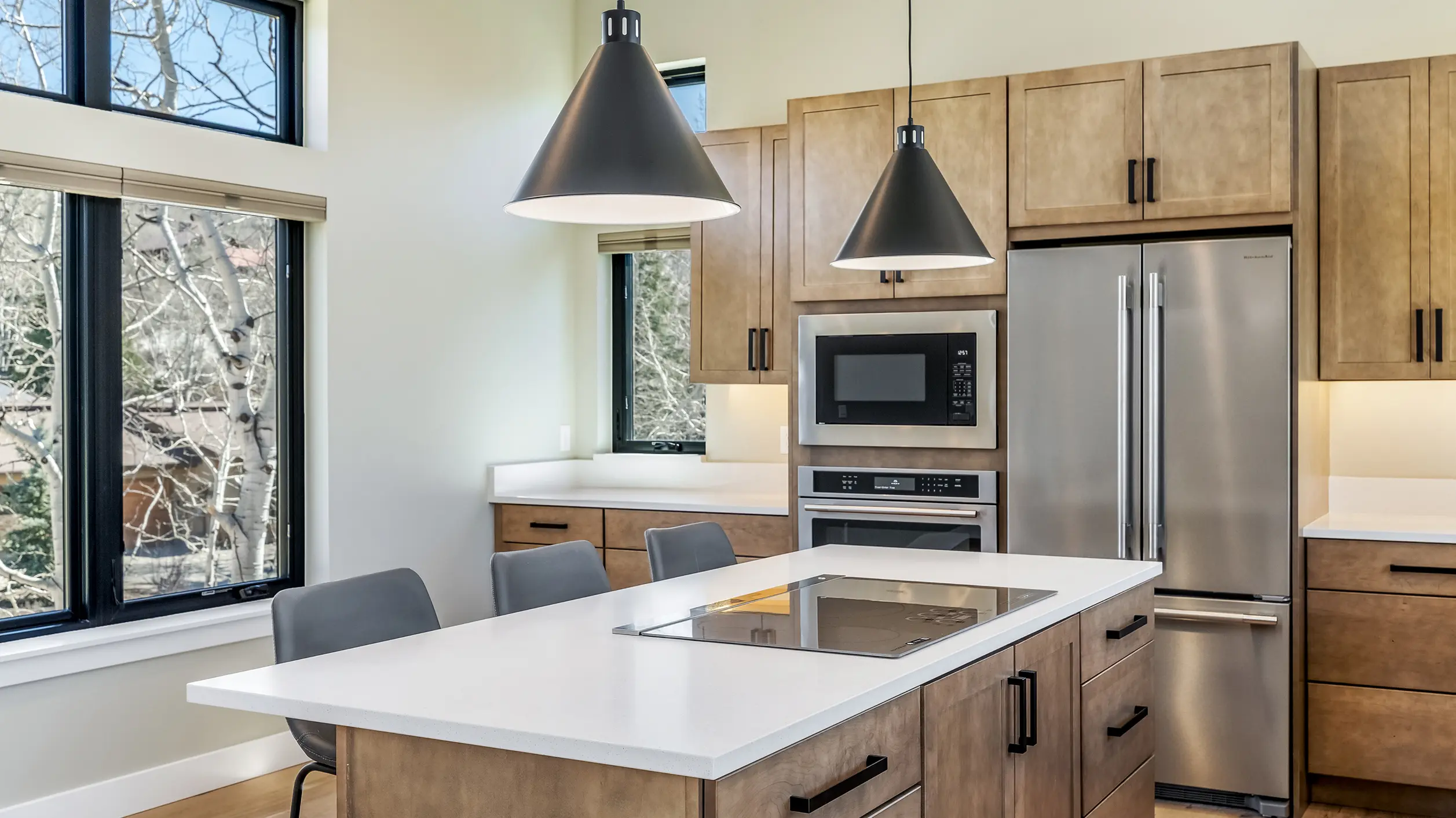 Telemark kitchen with warm wood cabinetry, white quartz island with black induction cooktop, black cone pendant lights, stainless appliances, and wall of windows